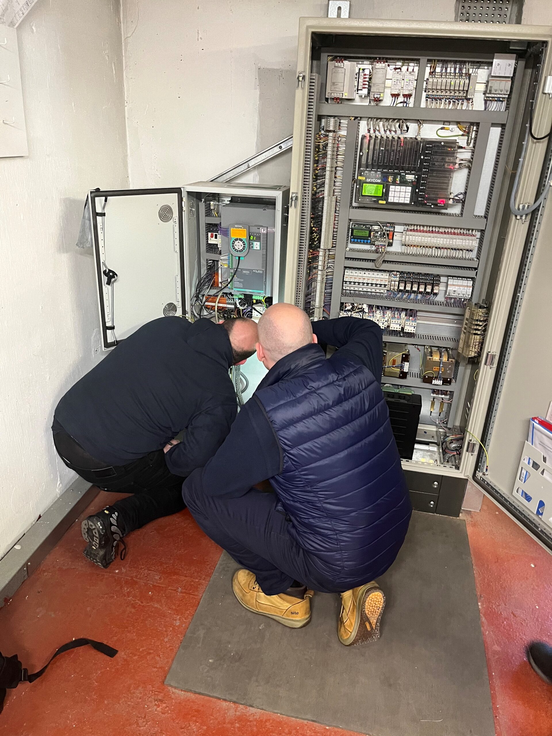 Two lift engineers crouching to look at lift controllers