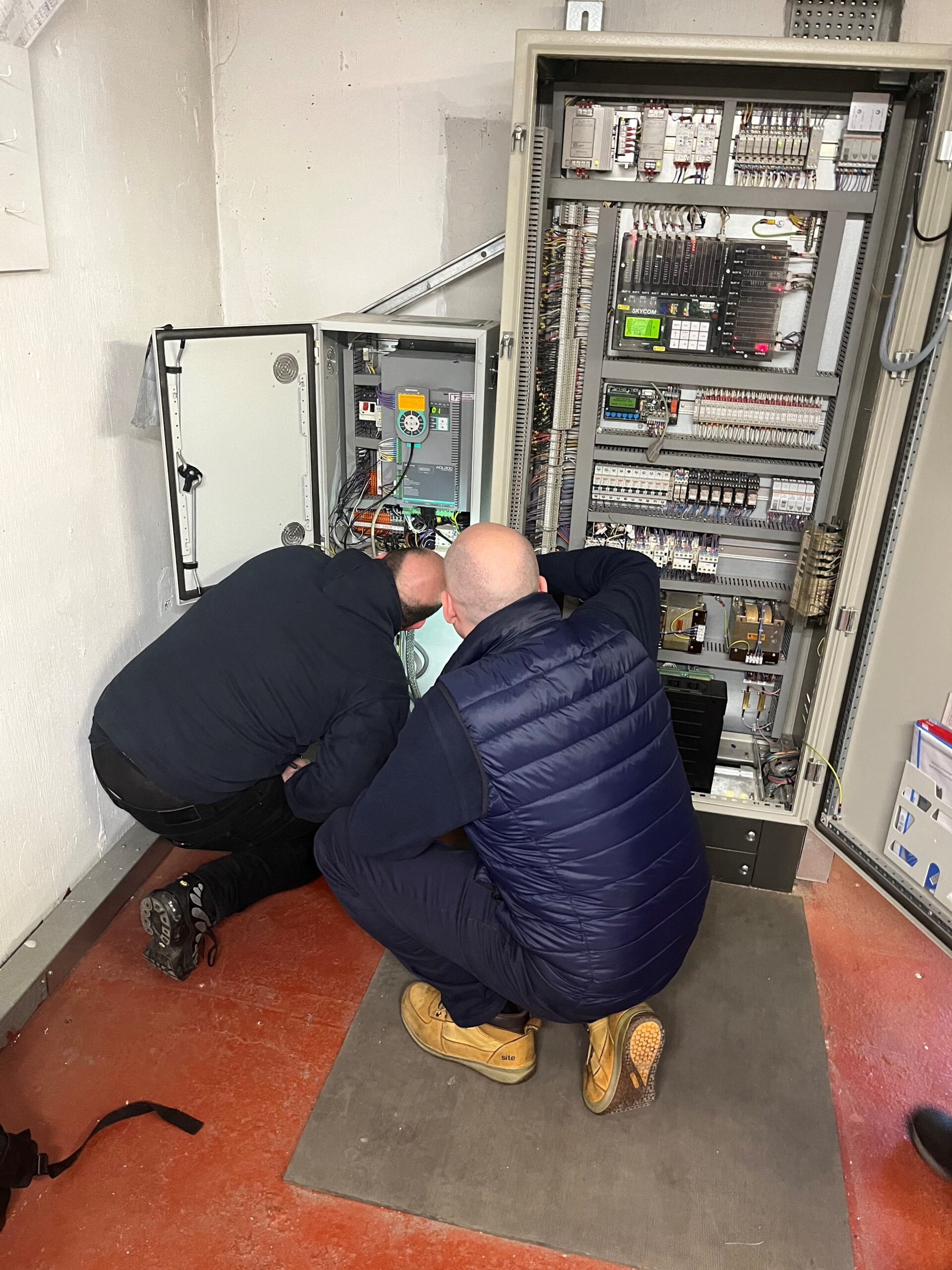 crouched-lift-controller Two lift engineers crouching to look at lift controllers